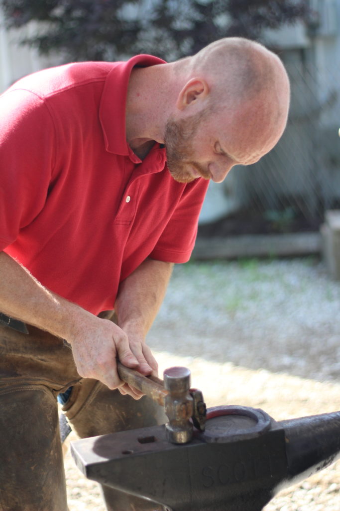 Photo of Cody
Bald man with red shirt standing at an anvil with a hammer and horseshoe